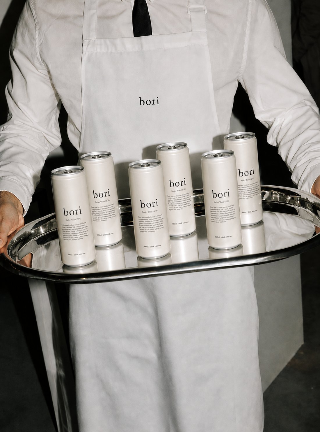 A server in a bori apron carrying a silver tray of bori barley water cans