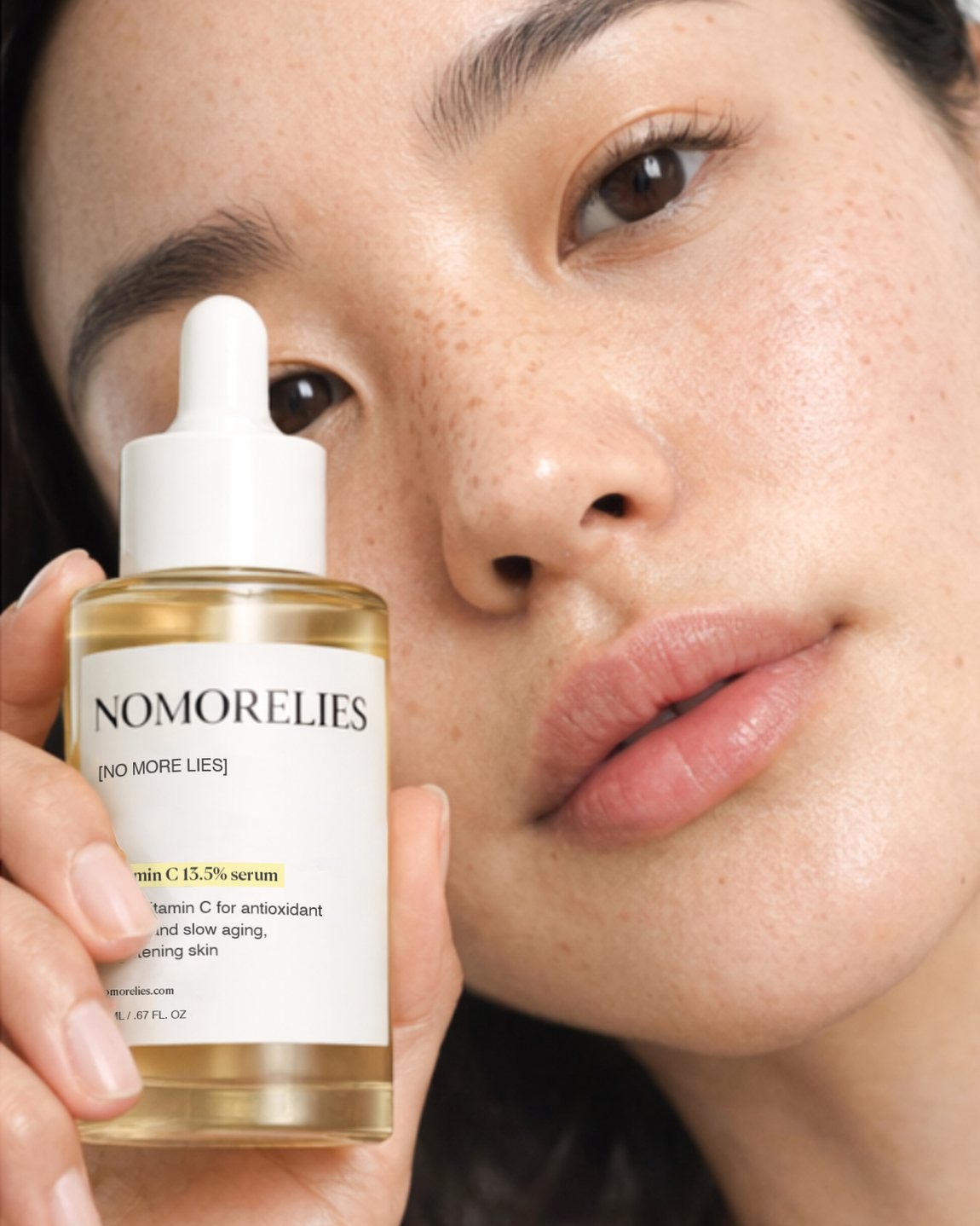 A close-up of a woman holding a Nomorelies vitamin C serum bottle beside her face