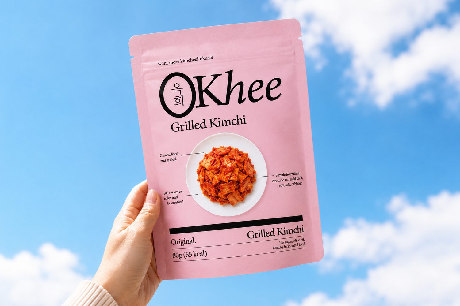 A hand holding a pink Okhee grilled kimchi pouch against a blue sky